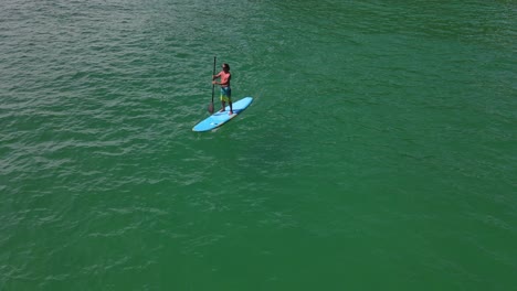 aerial drone bird's eye view of caucasian man exercising on a sup paddle board in turquoise tropical clear waters, with beach and coastline in thailand