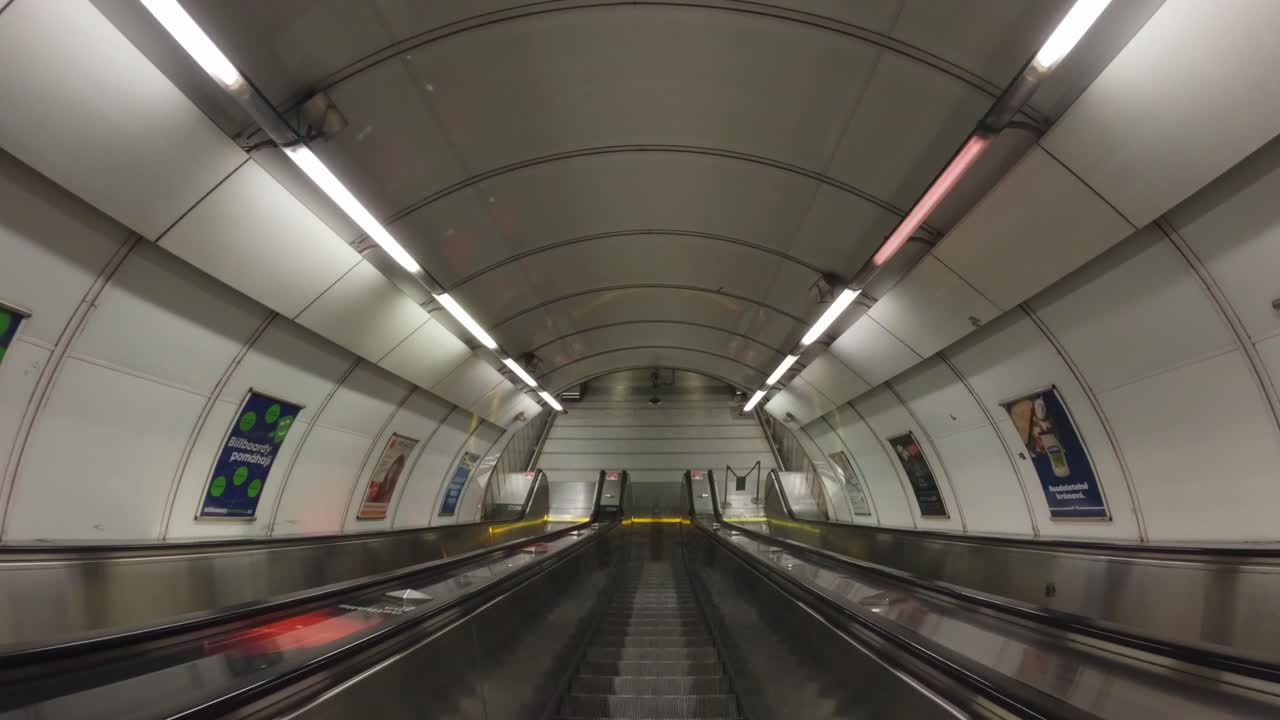 A long underground metro escalator with a curved ceiling and bright fluorescent lighting. Advertisements line the tunnel walls, adding a touch of urban character.