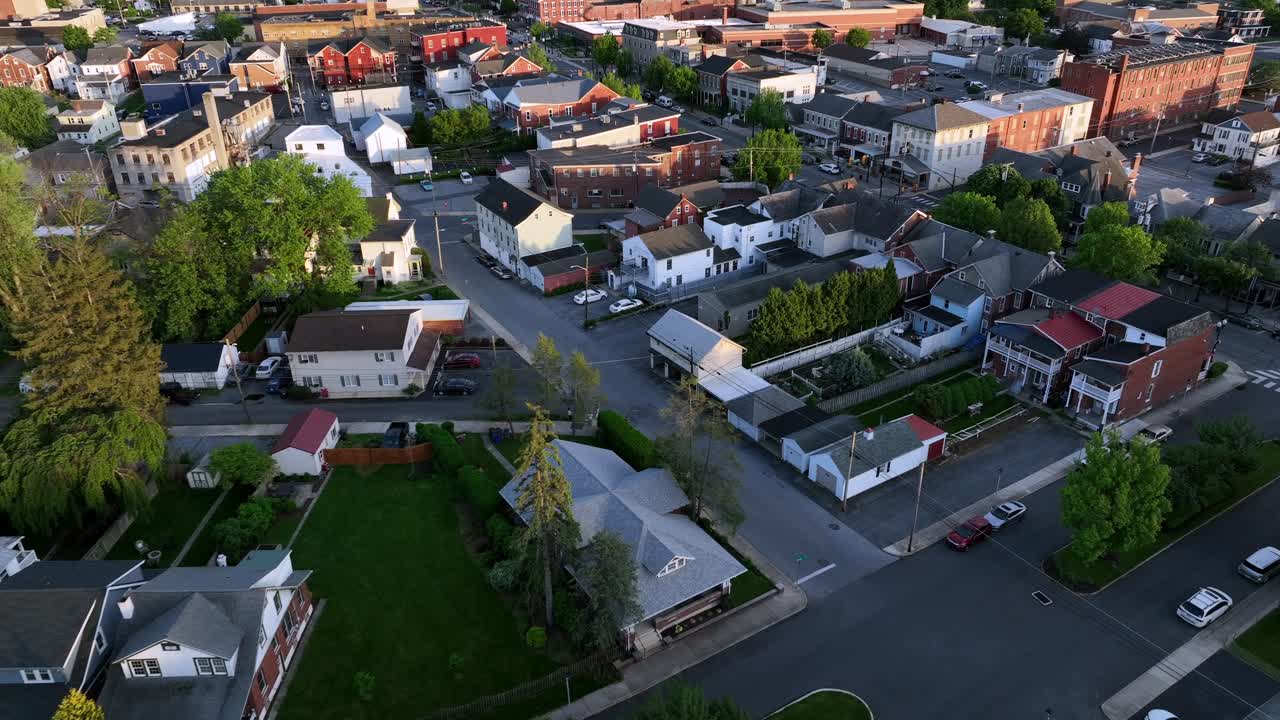Drone flight over american housing area with row of homes and driving cars on junction. Sunset time in historic town in springtime. Red brick buildings of downtown in distance. Top down.
