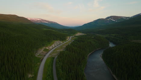 Mignight-sun-spreads-over-E6-highway-following-winding-river-below-dense-pine-tree-forest,-aerial-panoramic