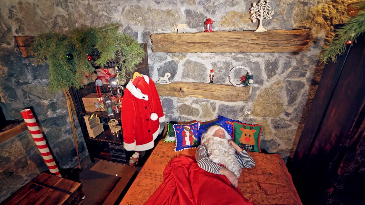 Santa Claus is sleeping in specially decorated room in his bed. He is holding his hand under his cheek. Christmas spirit concept close up video. Christmas eve. New Year is coming. Video from above.