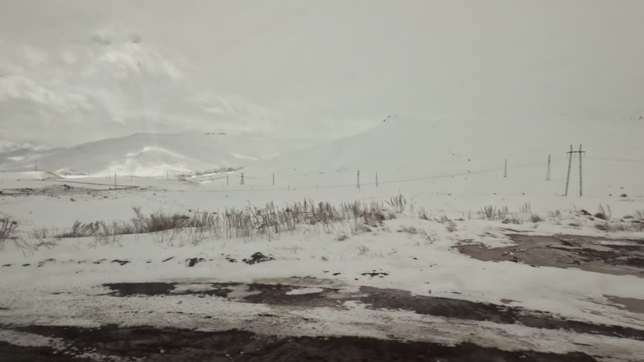 A wide view of a snowy landscape shows power lines stretching across the scene. A small building and an old structure appear in the distance among the snow