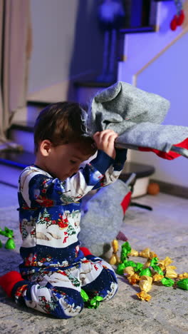 dark-haired baby boy in winter pajama sits near christmas tree. lovely boy gets some candies from the stocking. vertical video