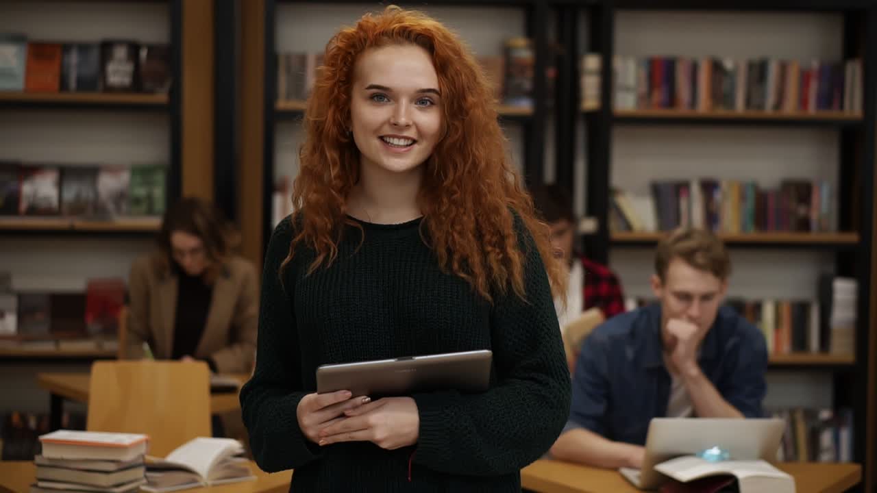 Portrait of an attractive long red haired european girl student standing in high school library smiling smiling looking at camera while holding a tablet. Education, literature and people concept. Classmates on blurred background