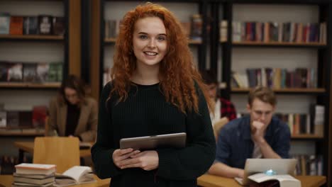 Portrait-of-an-attractive-long-red-haired-european-girl-student-standing-in-high-school-library-smiling-smiling-looking-at-camera-while-holding-a-tablet.-Education,-literature-and-people-concept.-Classmates-on-blurred-background