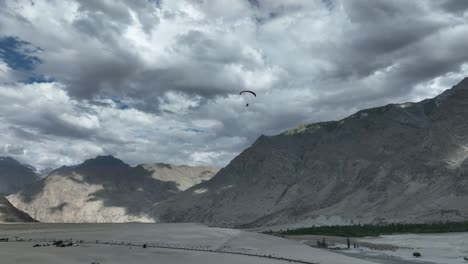 Drone-shot-of-the-sarfaranga-desert-with-a-view-of-high-mountains-and-peolpe-with-Paraglider