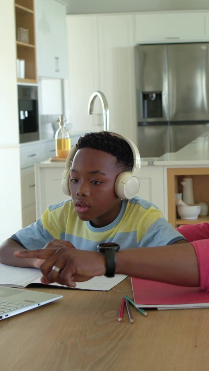Vertical video: Entering dad pointing at laptop, guiding son in striped tee in kitchen doing work