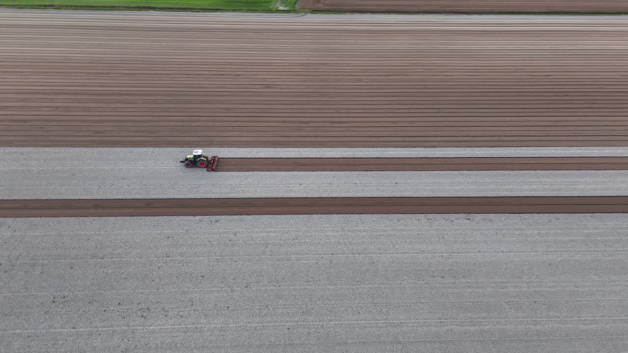 tractor pulling a harrow in slow motion, drawing deep brown seed lines beside bleached soil furrows across an expansive field as warm dusk light fades into evening slow motion drone shot