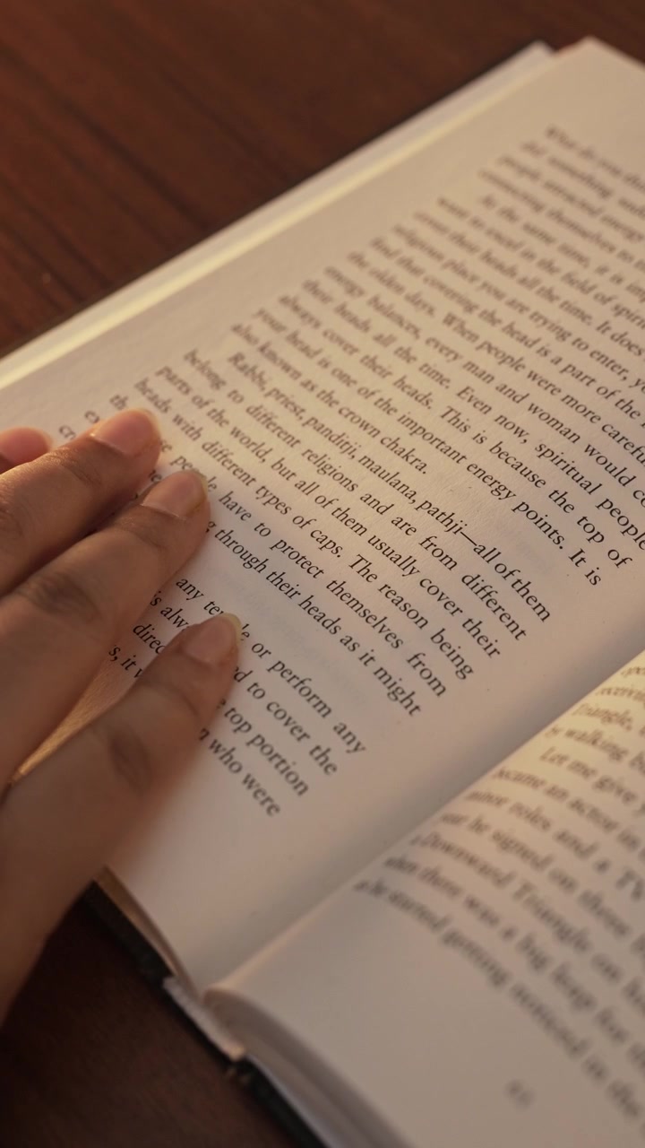 Close-up of a woman's hand gently pressing and smoothing an open book page while reading, with clearly visible printed text, warm wooden table surface, and warm light illuminating.