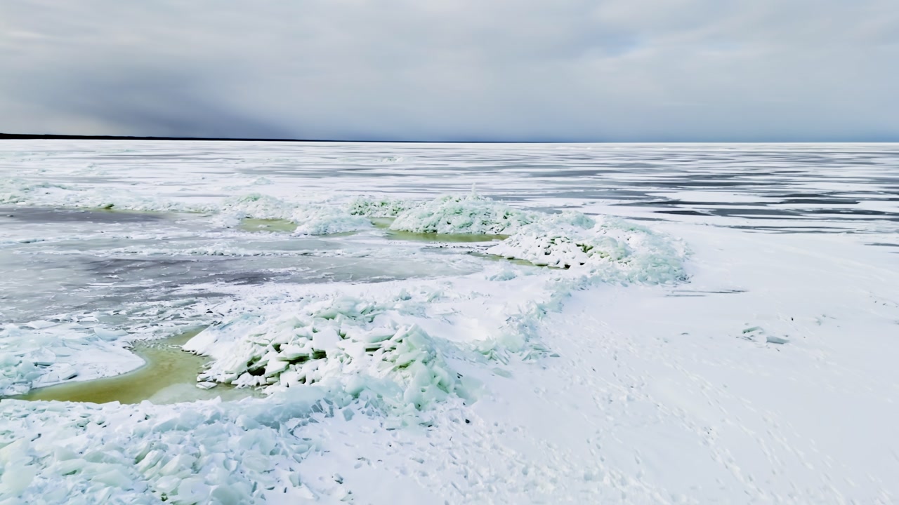 Wide drone view of frozen sea ice ridges with a small handmade ice tower, snow-covered surface, and dramatic winter horizon under overcast sky.