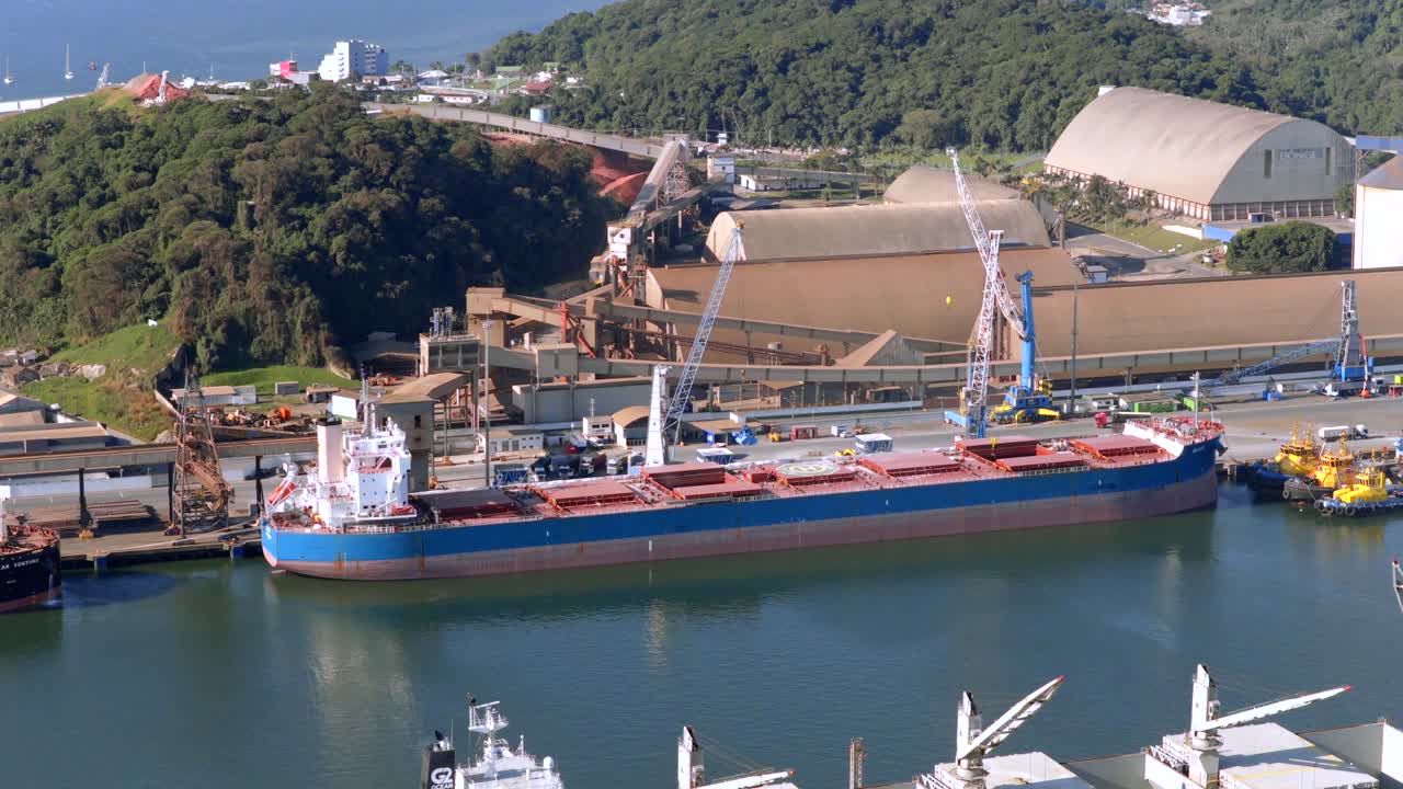 Slow, steady drone approaches large transport vessel docked at busy port, showcasing cranes and infrastructure during sunny afternoon
