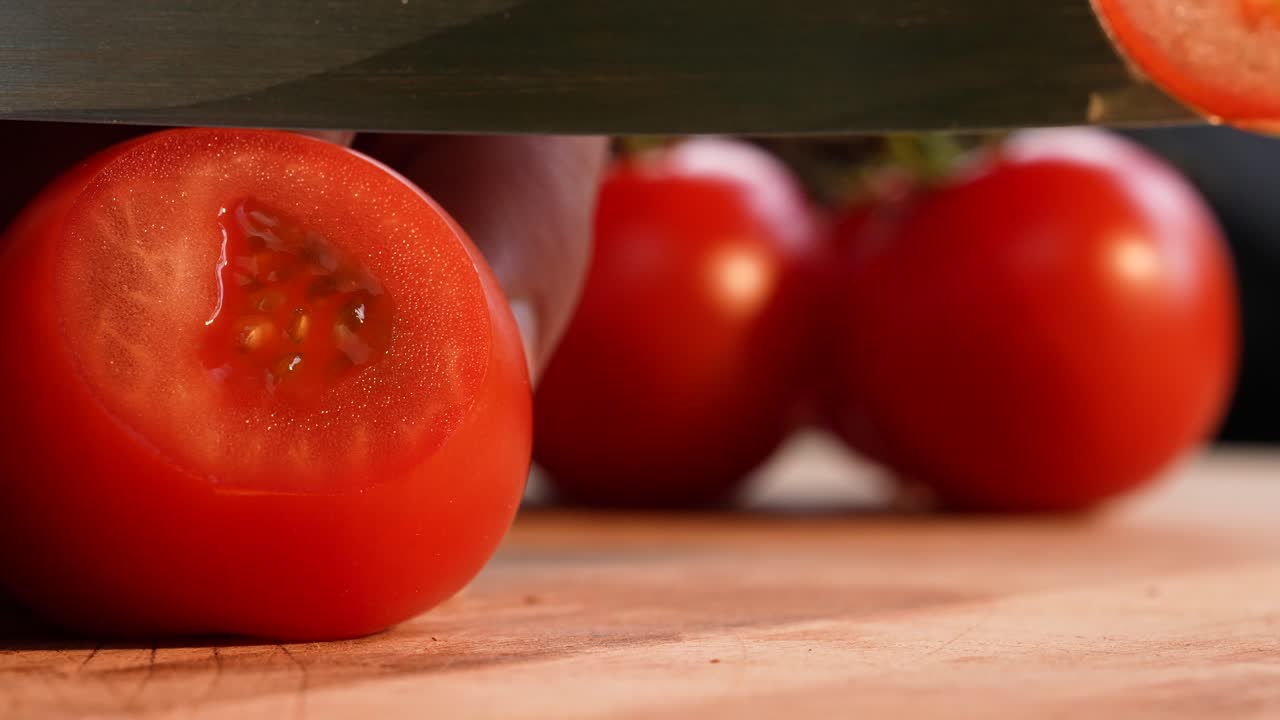 Landscape slow-motion close-up showing a tomato being cut into paper-thin slices with a razor-sharp blade. The smooth, controlled movement highlights precision and the delicate texture of the fruit
