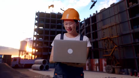 trabajadora asiática con gafas de protección y casco de seguridad usando una computadora portátil y con dolor de cabeza mientras está de pie en el sitio de construcción