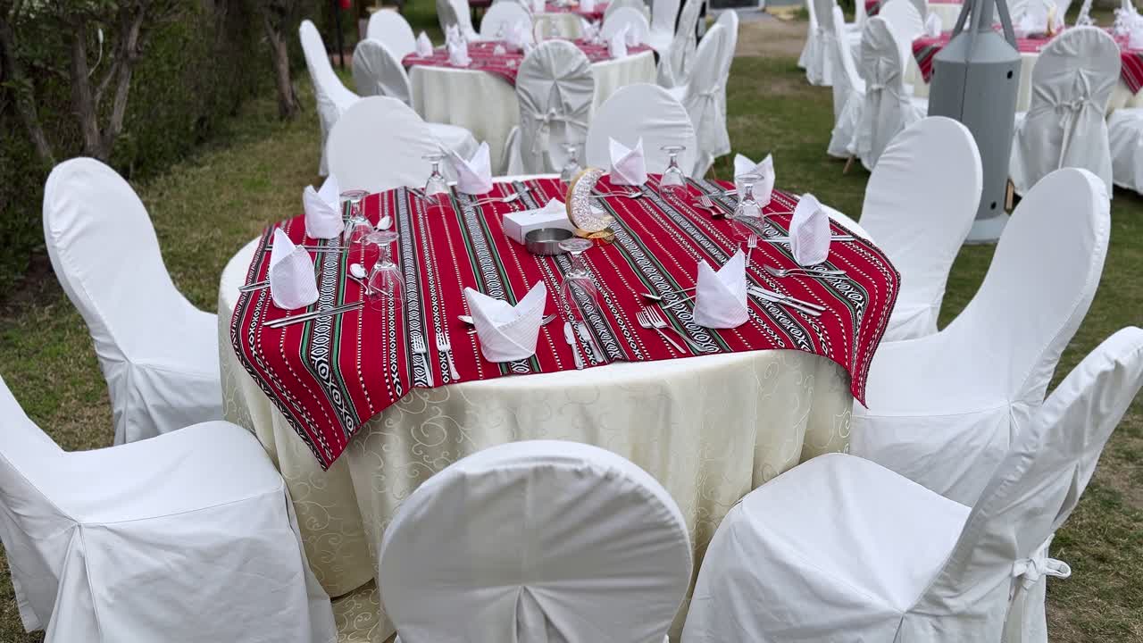 Bredt utsyn over et hagebankettoppsett med runde bord med røde tradisjonelle duker, hvite stoltrekk, glassvarer og halvmåneornamenter arrangert for en festlig iftar-feiring