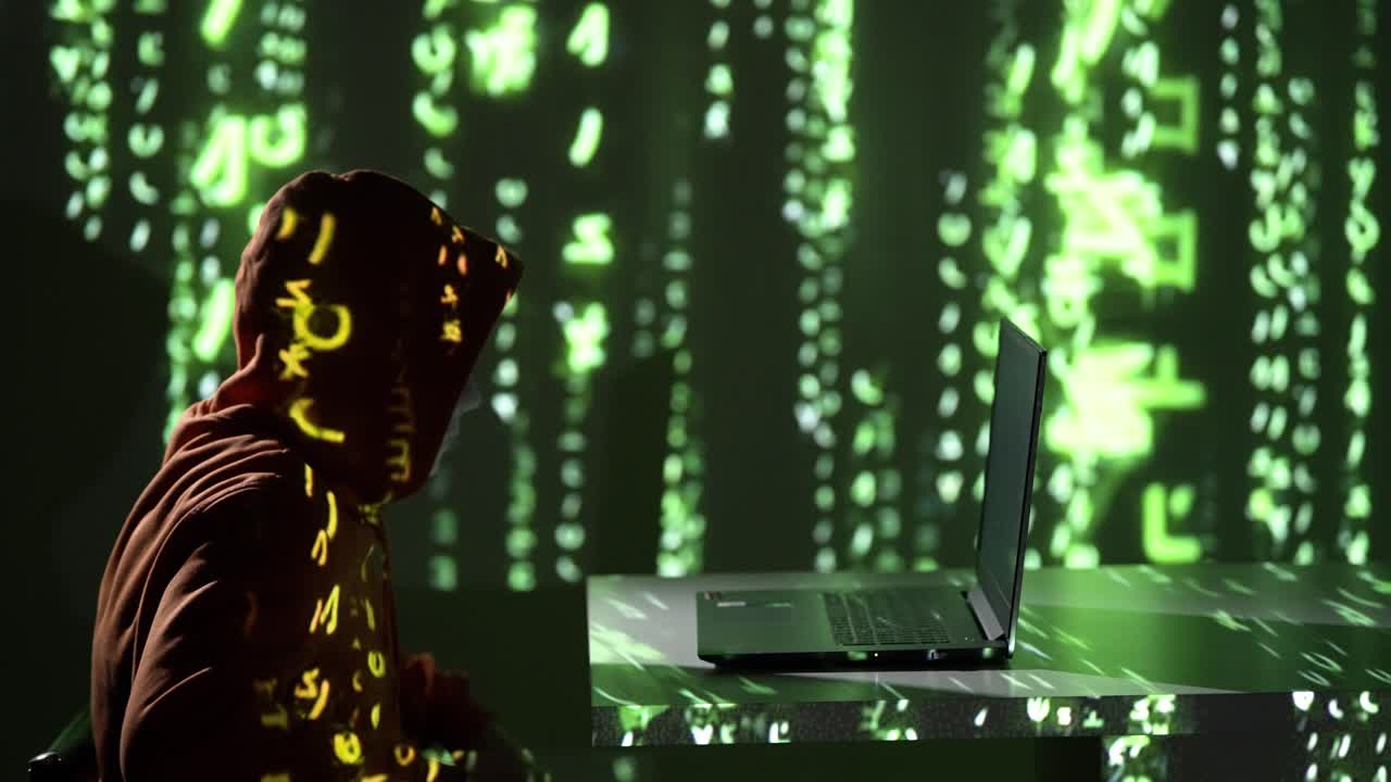 Hacker in hoodie leans over laptop as green code streams across the scene. A 4K slow-motion visual symbolizing cyber stress, hacking, and data warfare in a dark digital environment