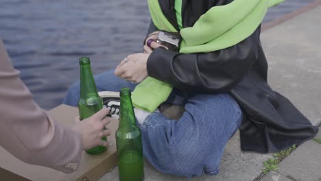 mujer joven con gorra negra y chaqueta de cuero sosteniendo una cámara junto a botellas de cerveza en la orilla del río