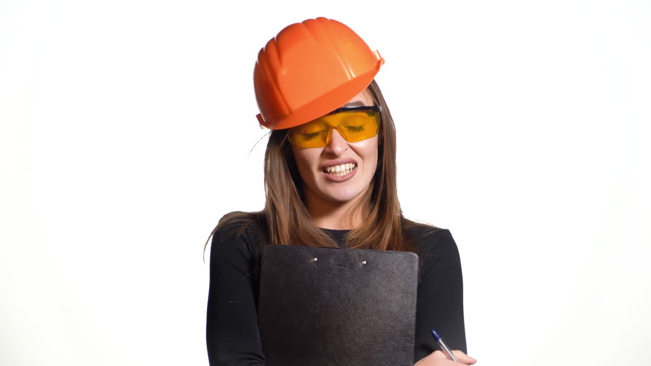 A beautiful woman in an orange helmet and in protective glasses holds a black planchette and pen in her hands and talks about something on a white background.