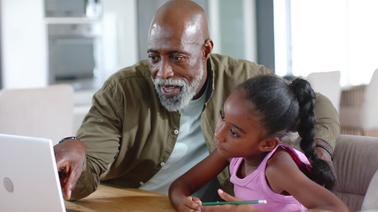 Senior African American man leaning pointing at laptop, guiding girl holding pencil at home table