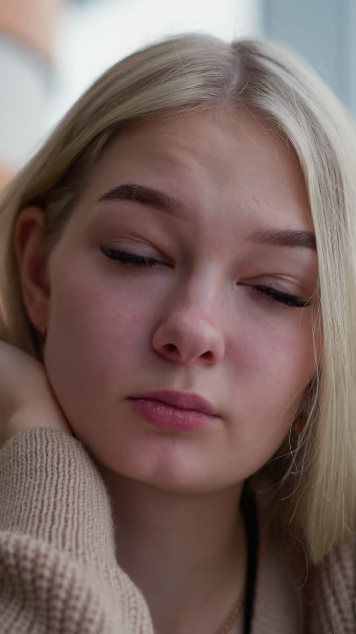 Young lady seated, resting chin on hand with pen in hand, looking tired and exhausted while glancing at busy urban setting through window, soft blur of city life outside
