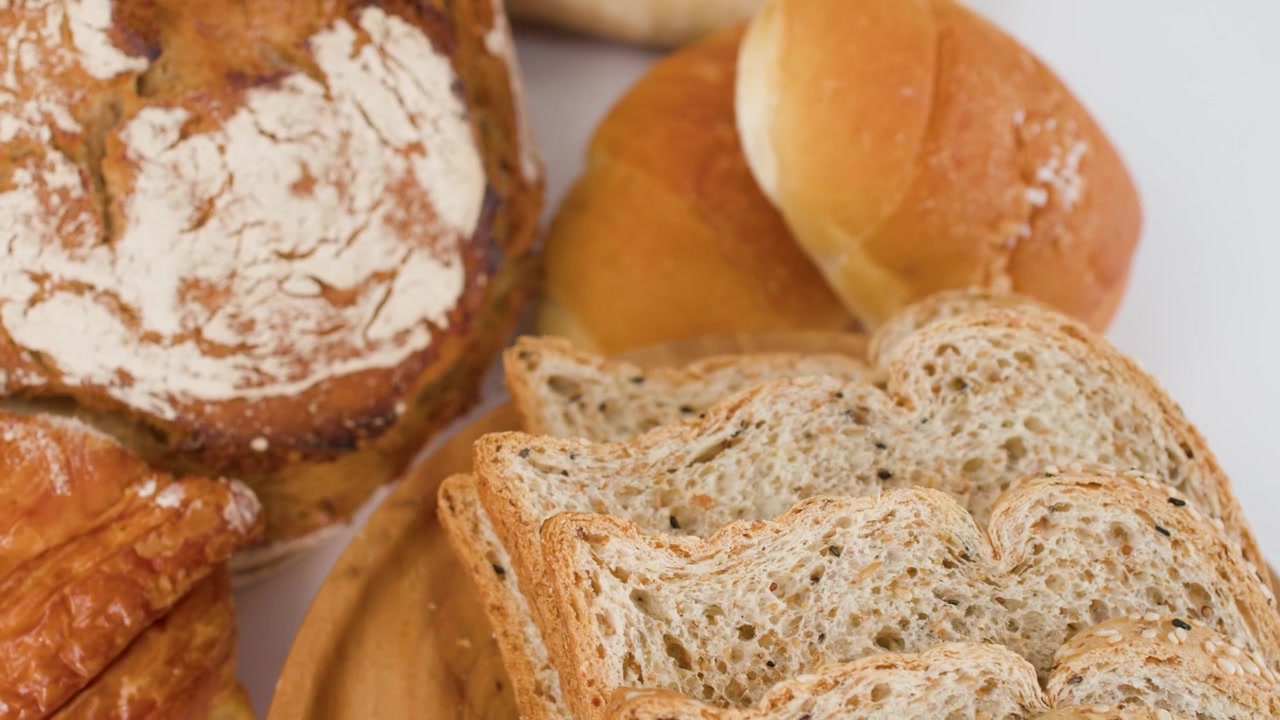 Vista ravvicinata rotante di pane a lievitazione naturale, croissant e pane multicereale affettato su un piatto di legno