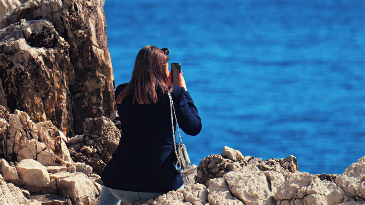 Woman sitting on the rugged rocks, on the shore, filming the blue sea with her phone