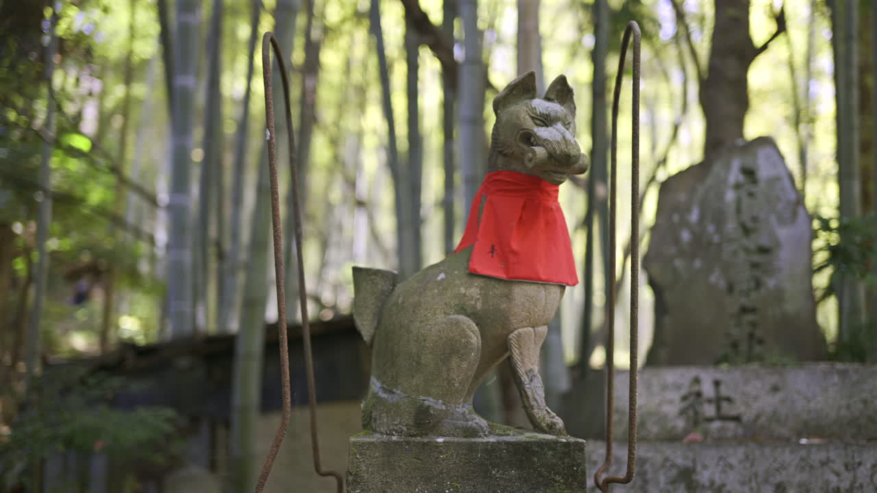 A stone fox statue adorned with a vibrant red scarf stands guard in a tranquil bamboo grove. Fushimi Inari, Kyoto, Japan