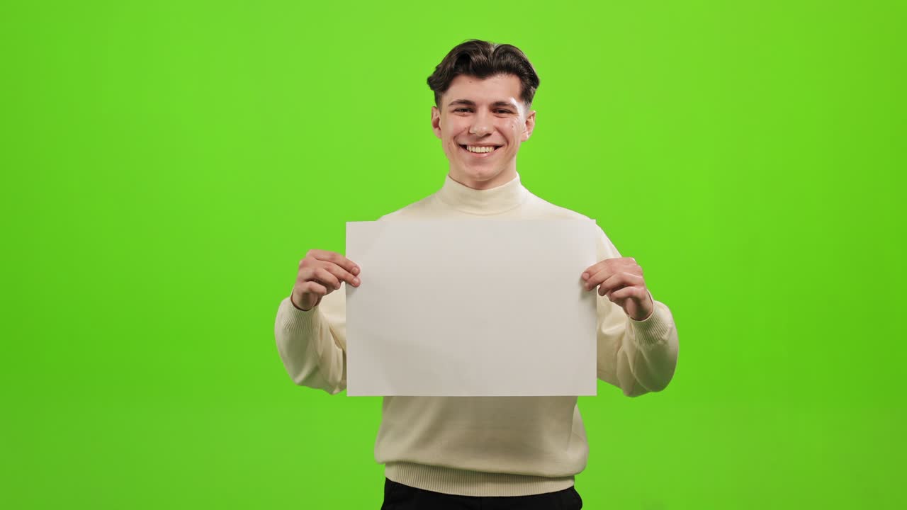 el hombre está de pie y mirando a la cámara. está levantando un papel blanco y sonriendo. está de pie sobre un fondo verde. pantalla verde. 4k.