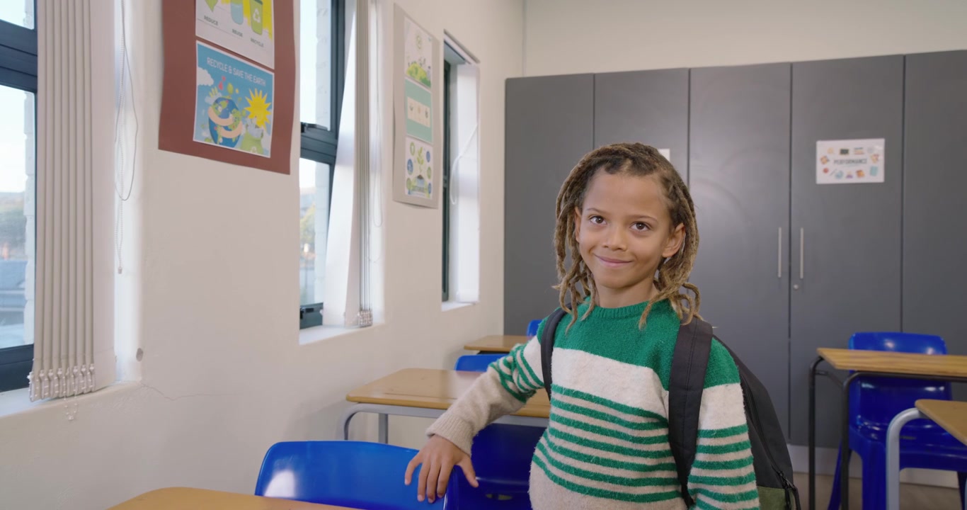 Ragazzo afroamericano con lo zaino in piedi al banco di scuola, la telecamera si stringe su di lui facendolo sorridere, con spazio per il testo