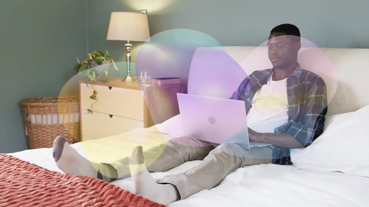 Man on bed using laptop for technology, glancing, typing while color overlay drifting across frame