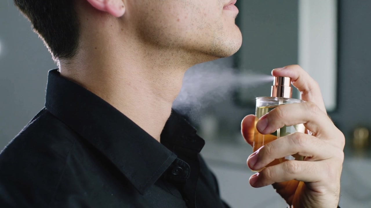 Spraying man raising bottle to neck, pressing nozzle, scenting skin in bathroom wearing black shirt