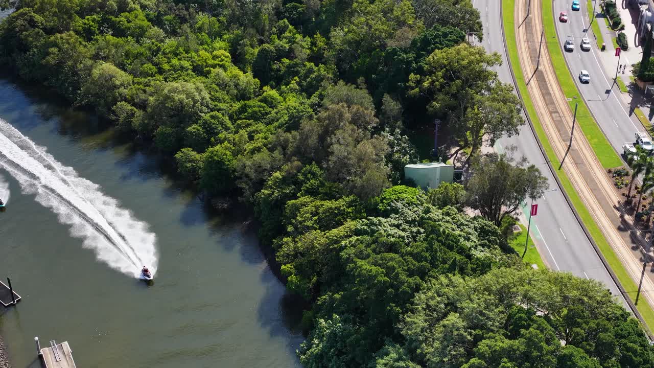 Aerial view of jet skis racing along a lush, tree-lined waterway near a busy highway in Gold Coast, Australia