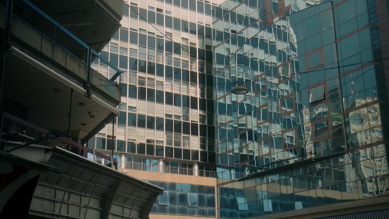 Close-up view of modern glass office building with reflective windows showcasing nearby structures. A blend of architectural styles is visible through reflections, adding depth to the urban scenery.