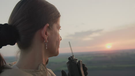 side profile of woman inside hot air balloon holding radio while gazing at glowing sunset horizon over vast green farmland and gentle clouds, capturing serene moment of communication
