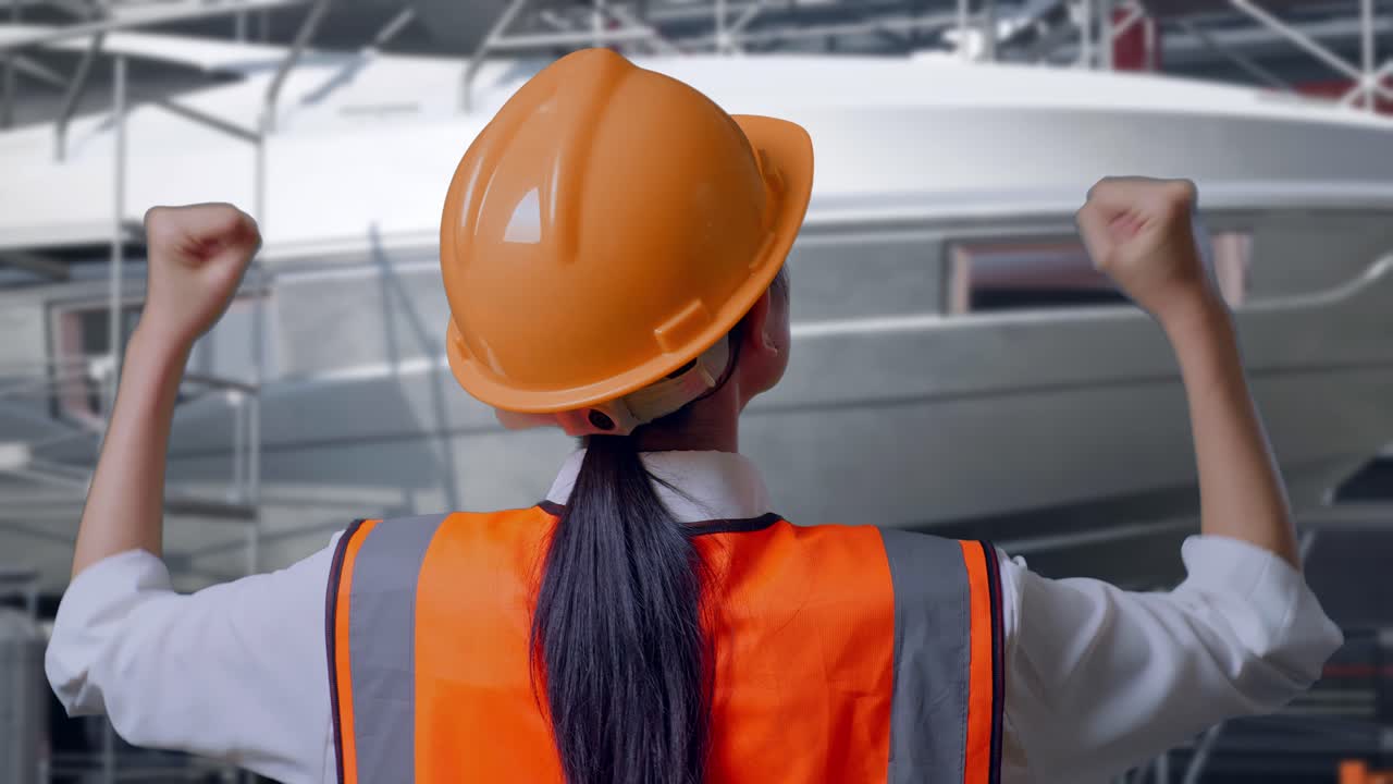 Primo piano da dietro di un'ingegnera con casco di sicurezza che alza le mani in segno di esultanza mentre lavora alla costruzione di uno yacht in un moderno cantiere navale
