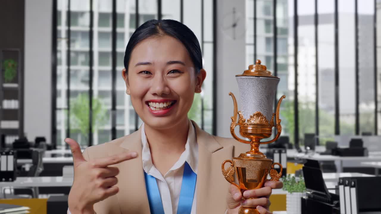 Close Up Of Asian Business Woman In A Suit With A Gold Medal Pointing To A Gold Trophy In Her Hand Being Proud Winning As The First Winner In The Office