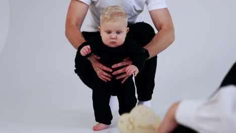 little baby in black suit walks supported by his dad. mom claps her hands to attract kid's attention. white backdrop.