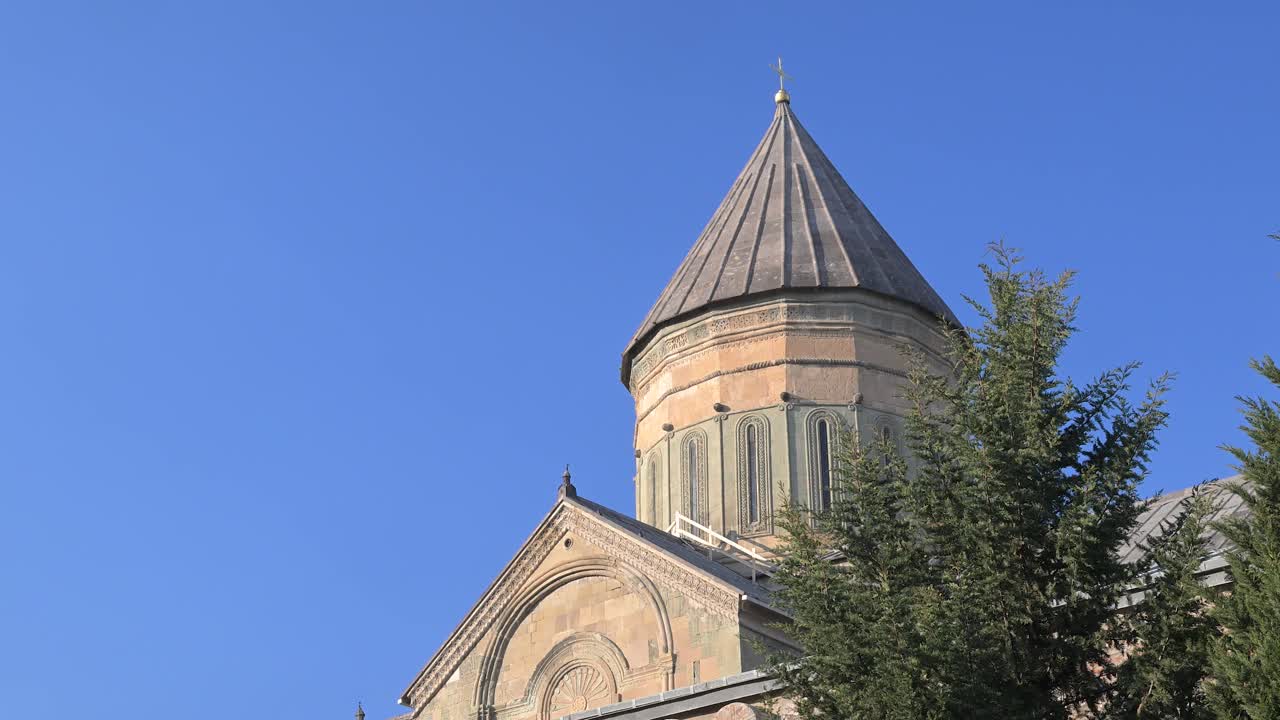 A view looking up towards Jvari Monastery shows the hilltop Georgian Orthodox church rising above the landscape with stone walls and early Christian design