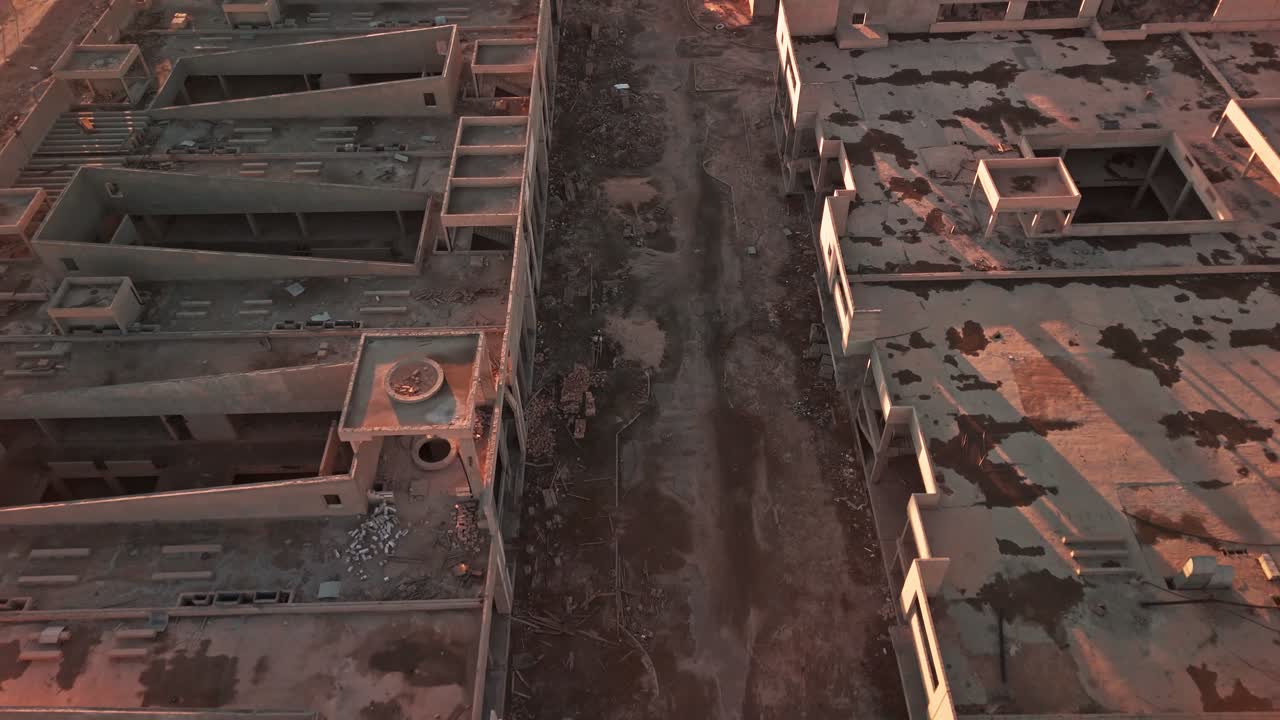 Vista aerea dall'alto di un cantiere abbandonato con edifici in cemento incompiuti, detriti e un corridoio sterrato; calda luce dell'ora d'oro che proietta lunghe ombre, atmosfera cruda di riqualificazione urbana