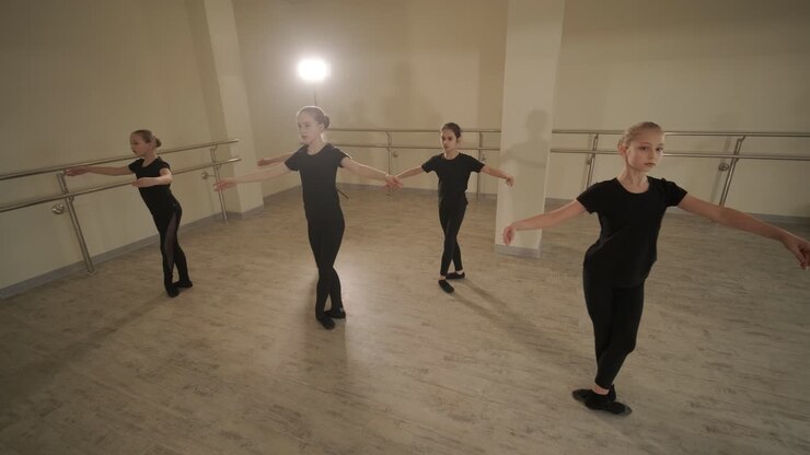 Un gruppo di giovani allievi di danza classica in abbigliamento da ballo nero che eseguono esercizi in un ampio studio di danza con pavimenti in legno e barre a muro. Espressioni concentrate e movimenti sincronizzati