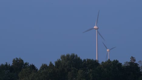 Granja-De-Turbinas-Eólicas-Que-Produce-Energía-Renovable-Para-Un-Mundo-Ecológico-Verde-En-La-Hermosa-Puesta-De-Sol,-Bosque-Verde-En-Primer-Plano,-Plano-Medio