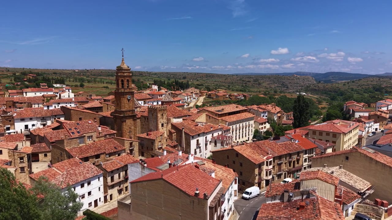 Vista panorámica de un pueblo medieval español con tejados de terracota, un campanario de iglesia histórico y campiña verde ondulada bajo un cielo azul