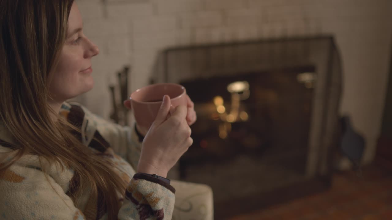 A cozy fall setting with a woman in a sweater enjoying a hot cup of tea in front of the fireplace. Over the shoulder shot at 60 Frames for slow motion