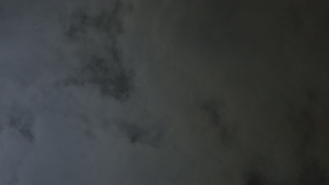 Bløde, mørke skyer bevæger sig langsomt på nattehimlen, svagt oplyst af måneskin. Et abstrakt og atmosfærisk visuelt element med en dyster tone.
