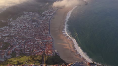 Blick-Auf-Die-Strandstadt-Nazare-In-Portugal-Mit-Niedrigen-Wolken-Bei-Sonnenaufgang,-Luftaufnahme