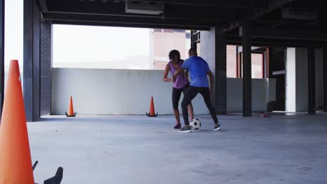 hombre y mujer afroamericanos jugando al fútbol en un edificio urbano vacío