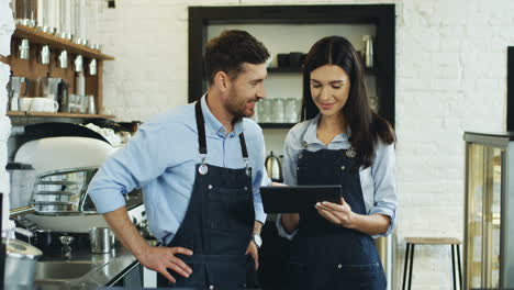 Junge,-Schöne-Kellnerin,-Die-Auf-Dem-Tablet-Gerät-Scrollt-Und-Tippt,-Und-Ihr-Männlicher-Kollege,-Der-In-Der-Nähe-Steht-Und-Den-Bildschirm-Im-Café-Beobachtet