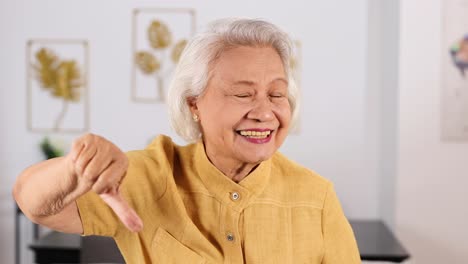 smiling asian grandmother in yellow shirt repeatedly gives thumbs down gesture in bright, modern room with soft natural lighting and steady camera framing