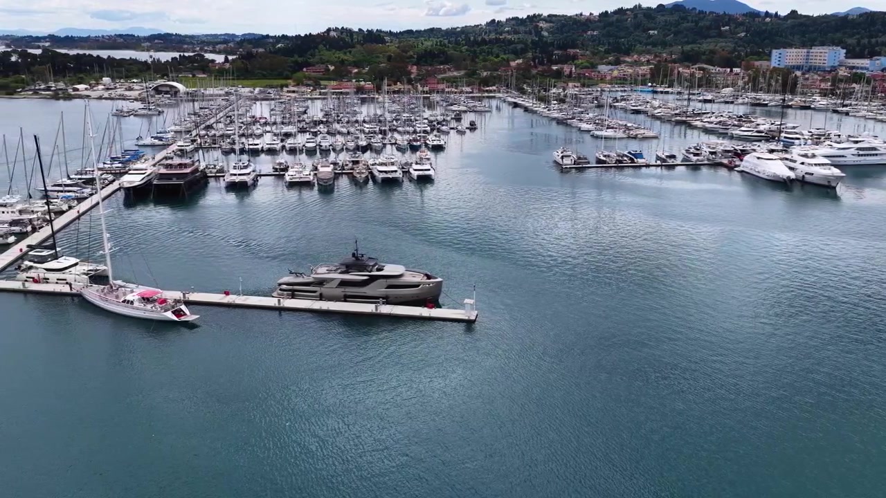 Vista aerea con drone della Marina di Gouvia a Corfù, Grecia, con yacht di lusso, barche a vela e catamarani ormeggiati in acque turchesi del Mediterraneo, sullo sfondo di lussureggianti colline verdi e di una suggestiva architettura costiera