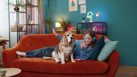child girl in wireless headphones relaxing listening music lying on couch at home near beagle dog
