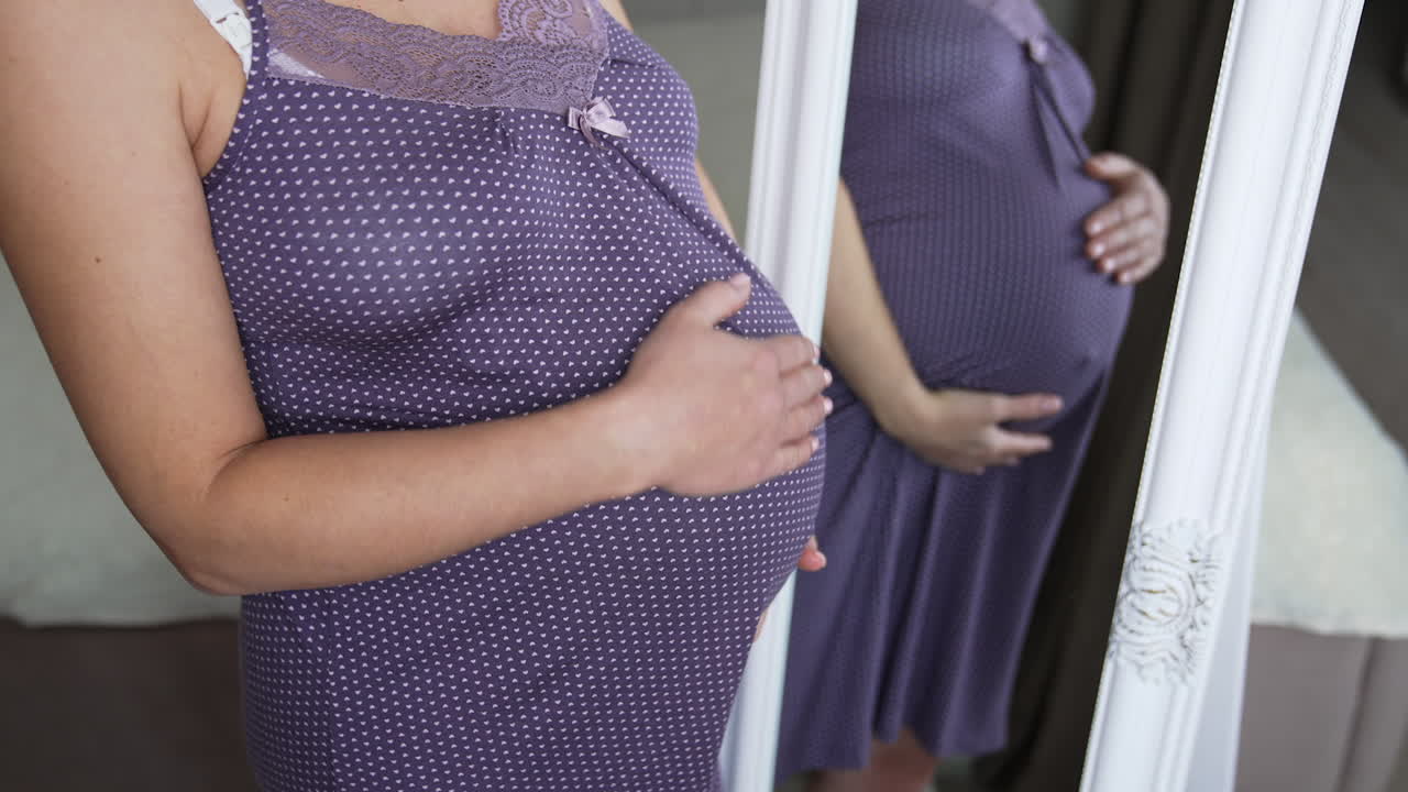 Woman waiting for child stands in a purple nightshirt in front of the mirror. Pregnant woman strokes her stomach with one hand reflecting in the mirror.
